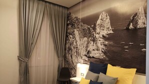 A hotel room with a bed, a chair, a desk, and a large wall mural of a coastal scene. - Beverello Rooms (Naples)