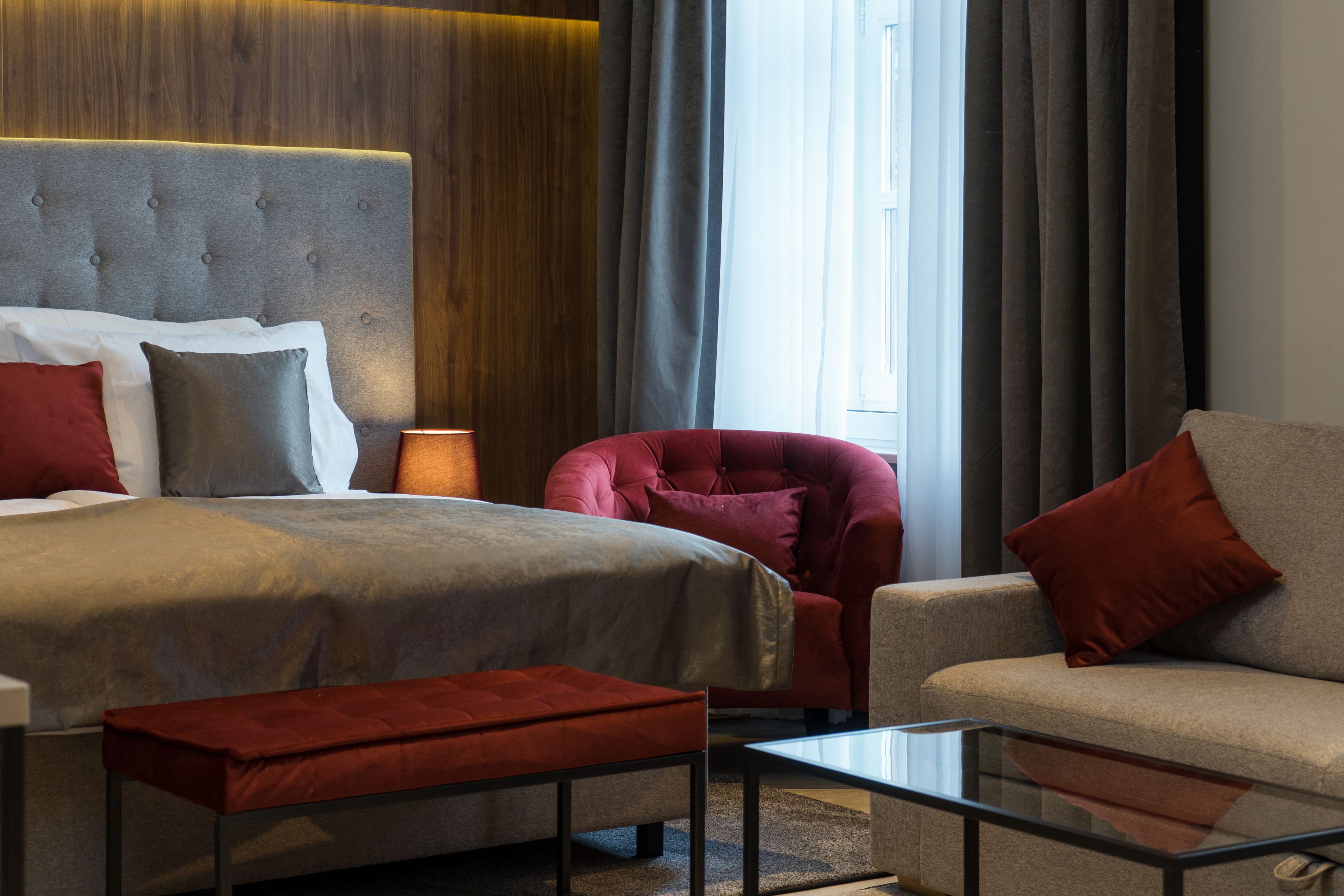 A modern hotel room with a large bed, a red armchair, a sofa, and a glass coffee table.