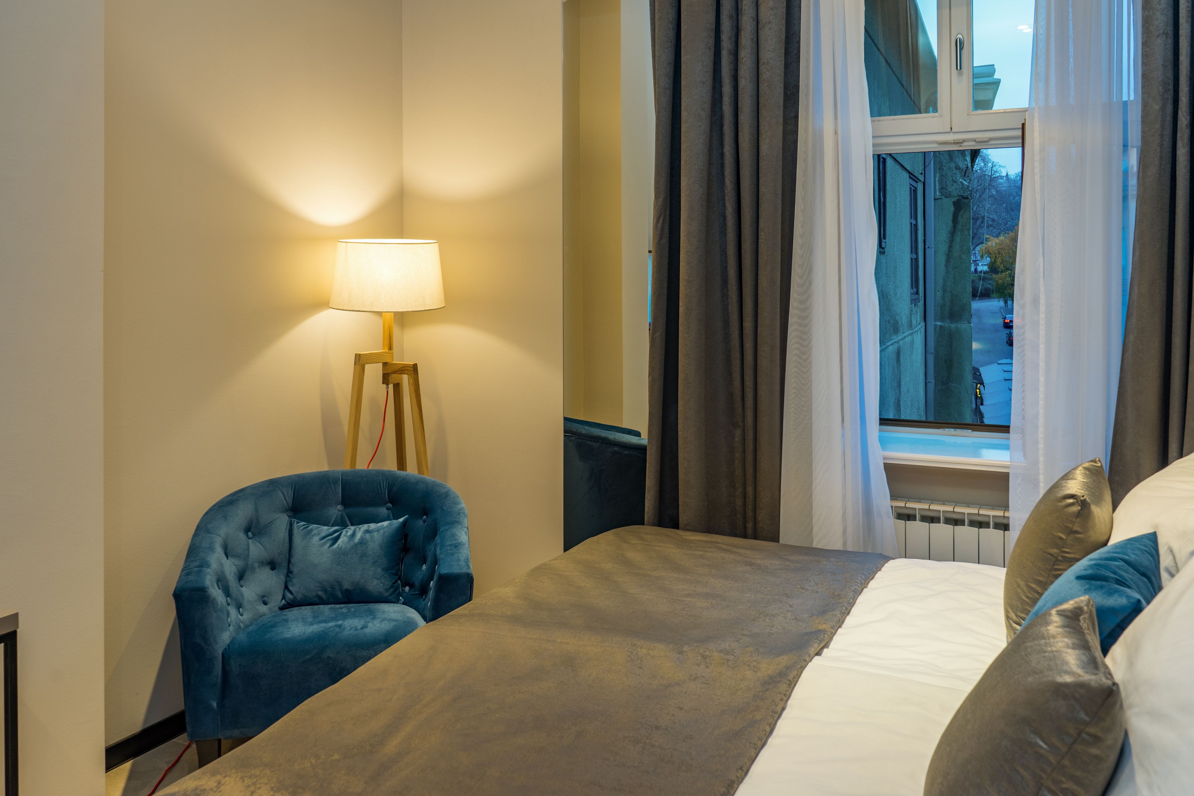 A hotel room with a bed, a blue armchair, a bedside table with a lamp, and a window with curtains.
