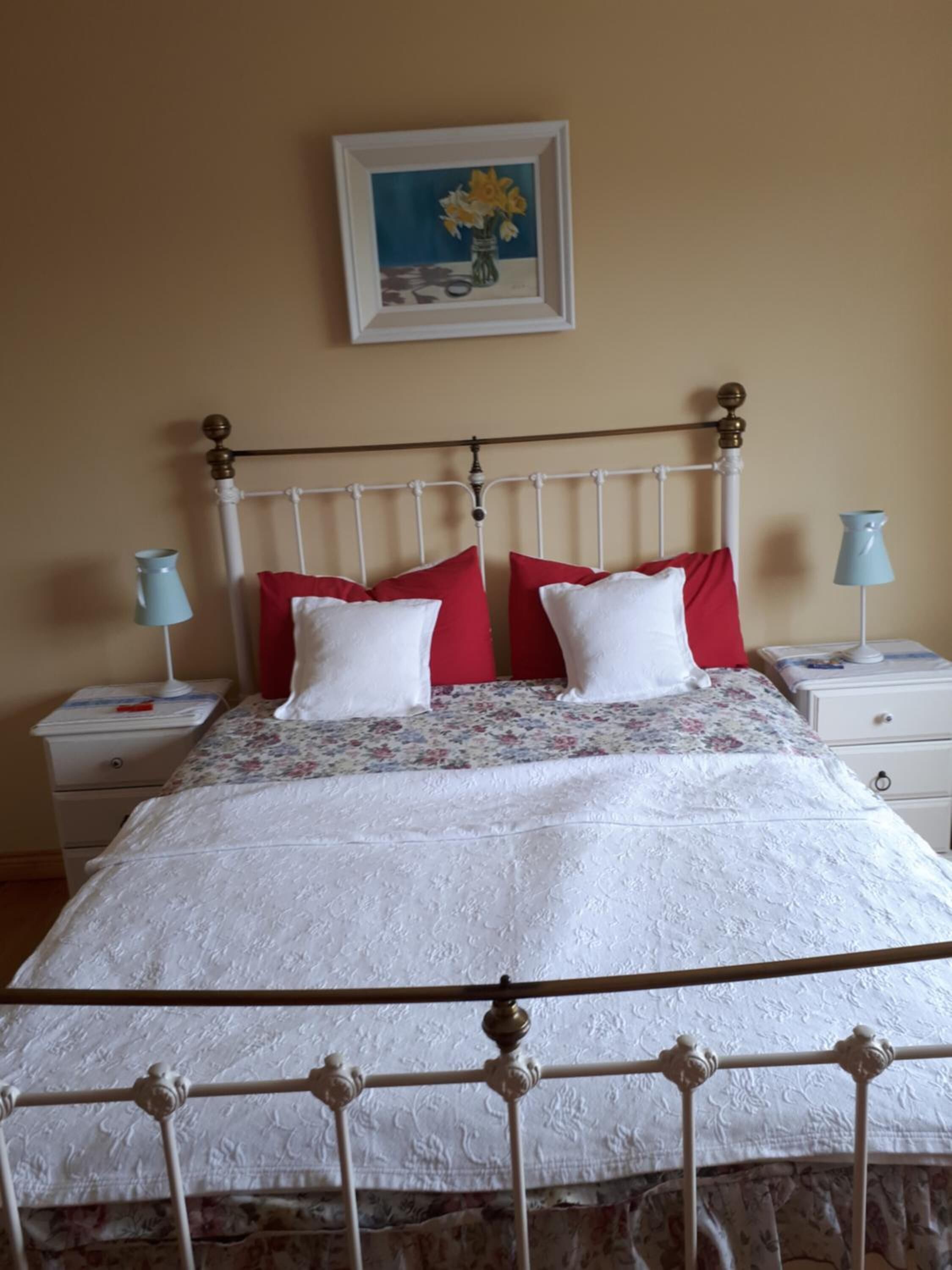 A neatly made bed with a white bedspread and red and white pillows, a metal headboard, and a framed painting above it.