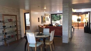 A dining area with a glass table, wooden chairs, and a red sofa. There is a stone pillar and a glass shelf with decorative items. - Casa Geribá (Búzios)