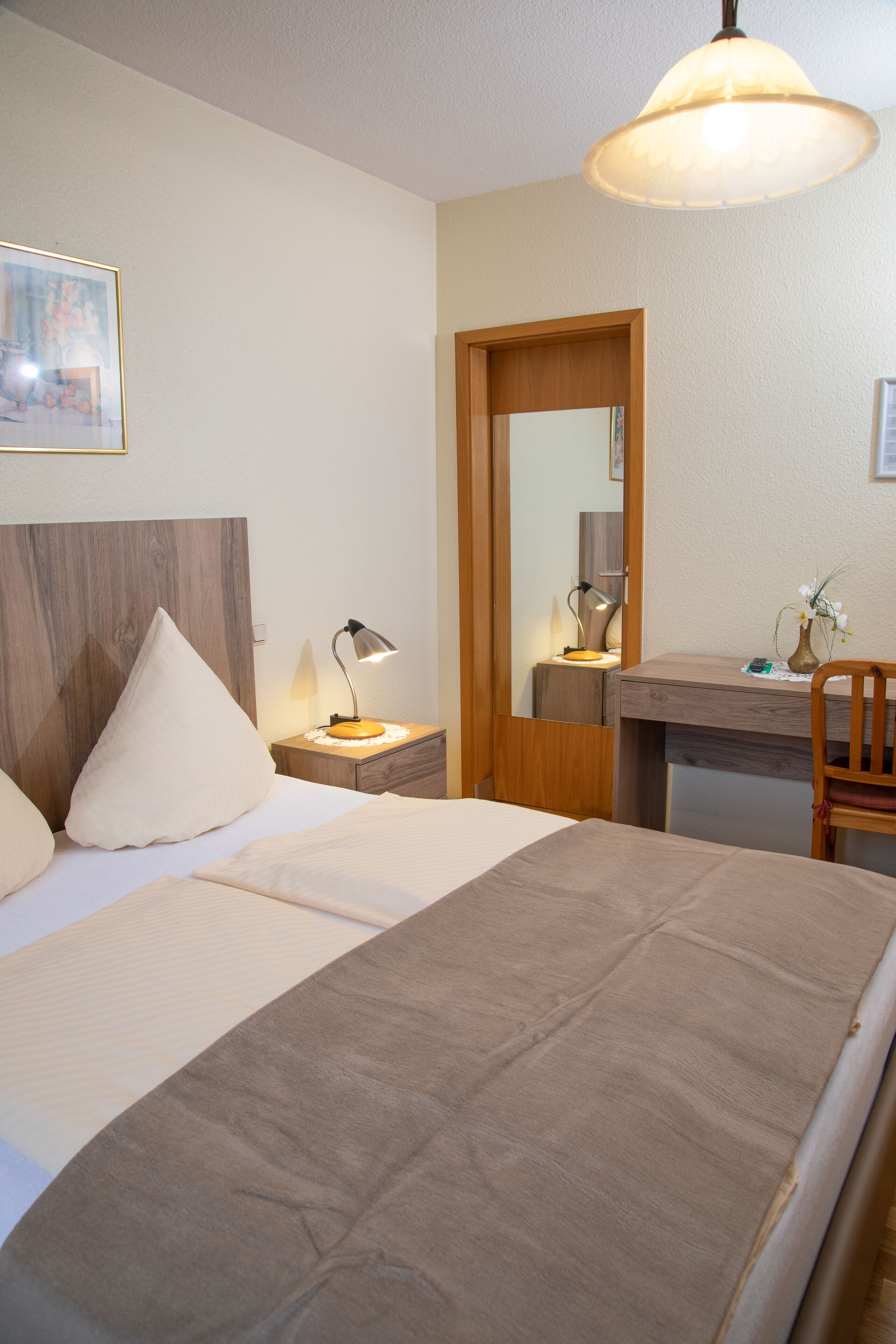 Una habitación de hotel ordenada con cama, mesita de noche, lámpara, silla y espejo.