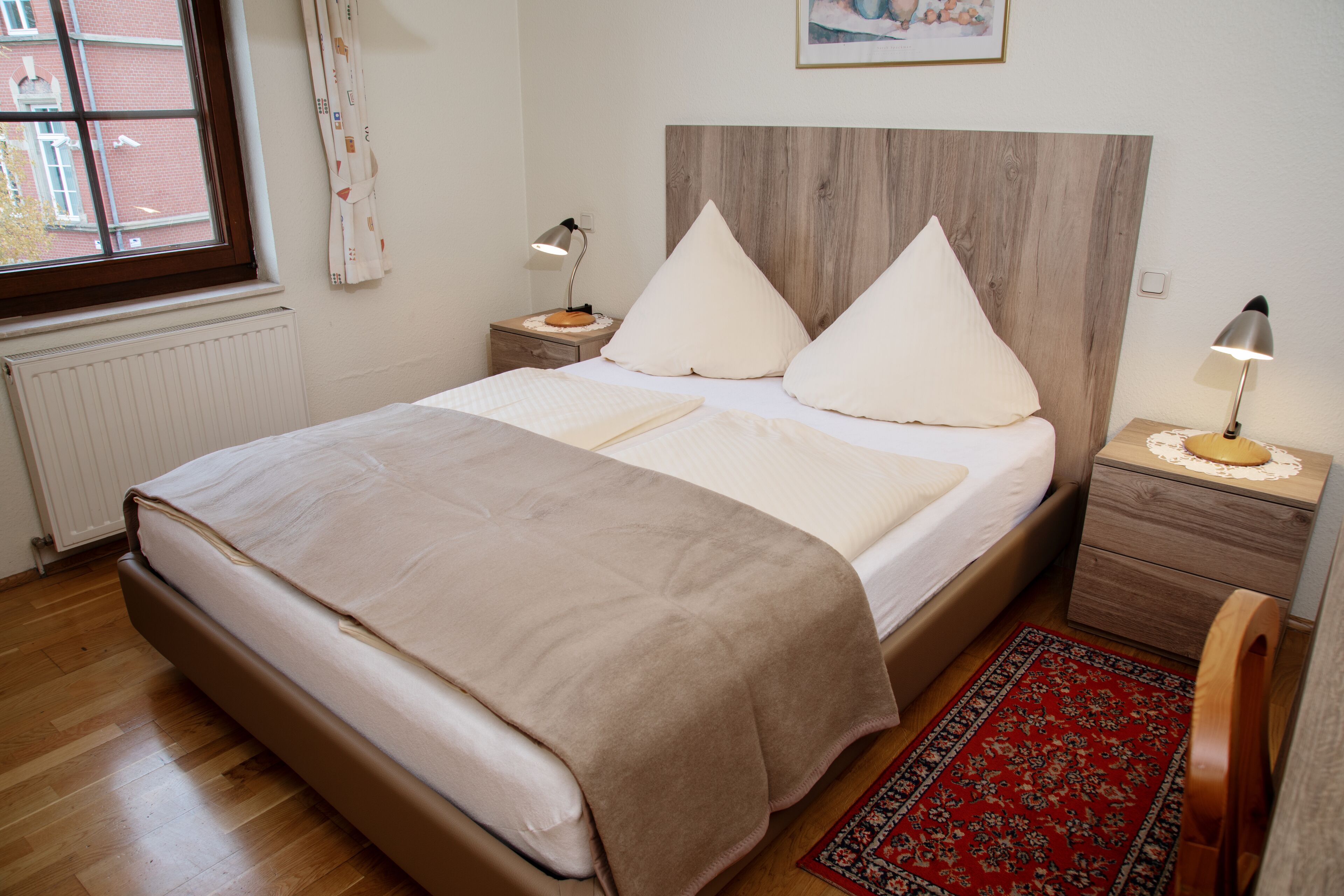 A bedroom with a wooden headboard, a large bed with two pillows, a bedside table with a lamp, and a red patterned rug.