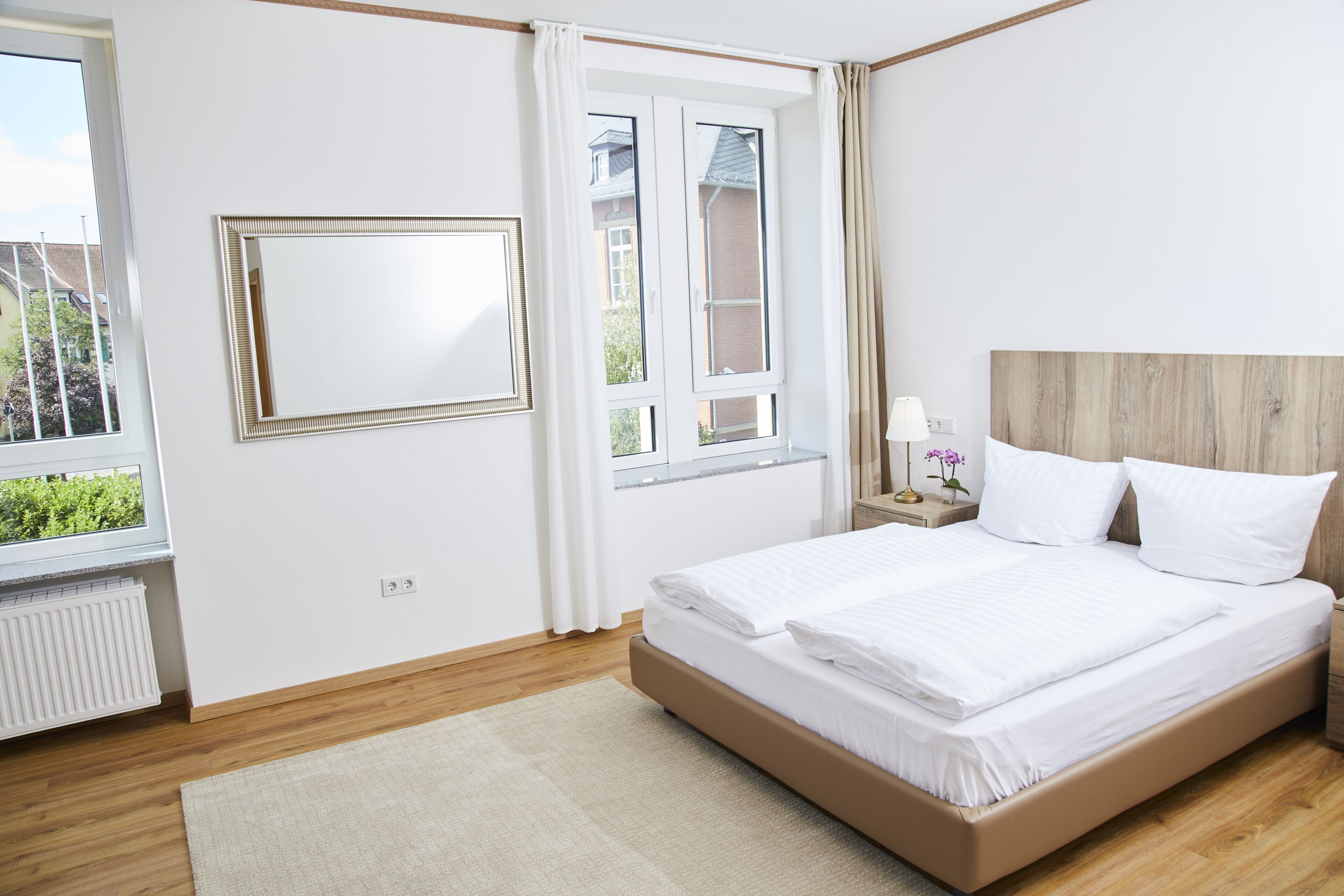 Un dormitorio con una cama grande, una ventana con cortinas, una mesita de noche con lámpara y un espejo en la pared.