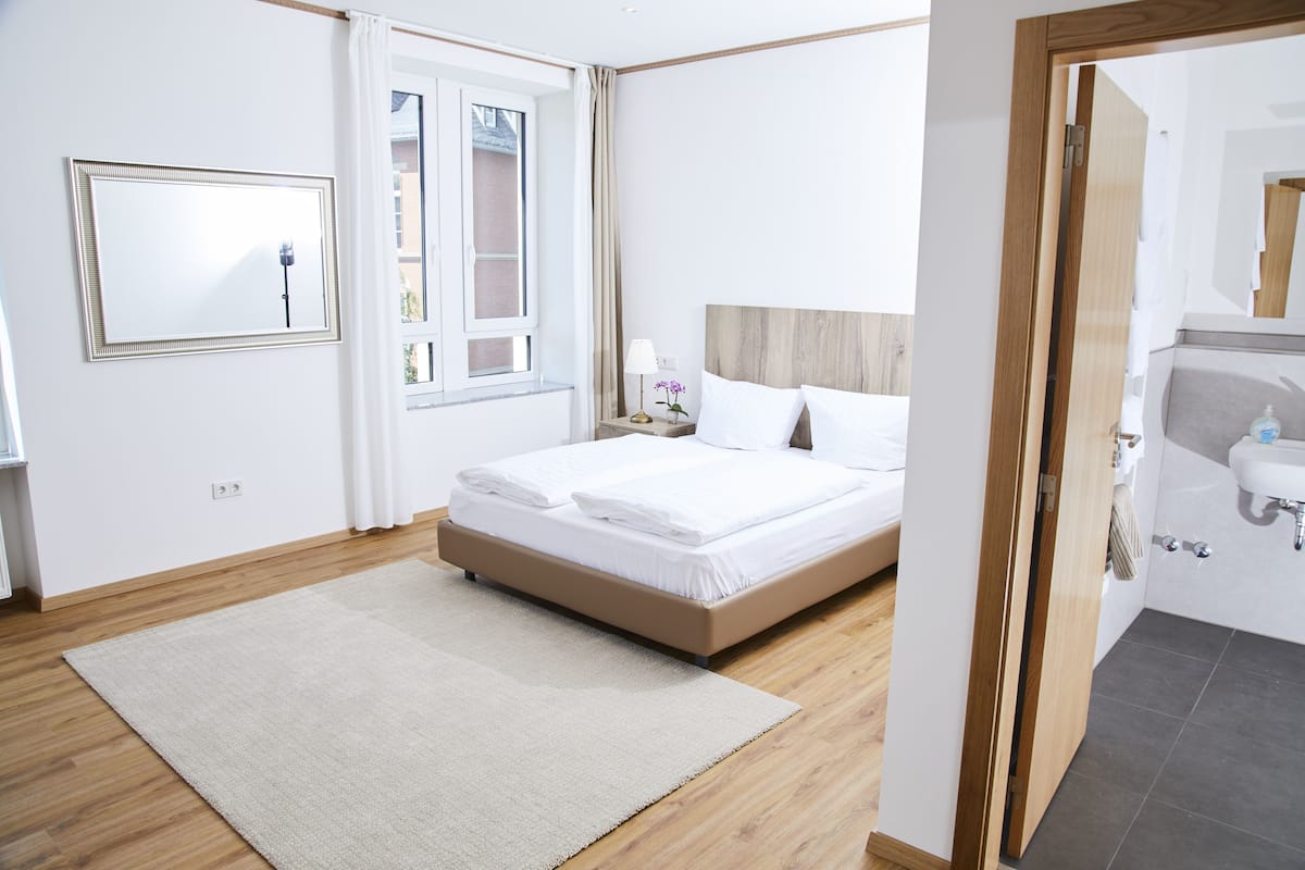A bedroom with a bed, a nightstand, a mirror, and a bathroom visible through an open door.