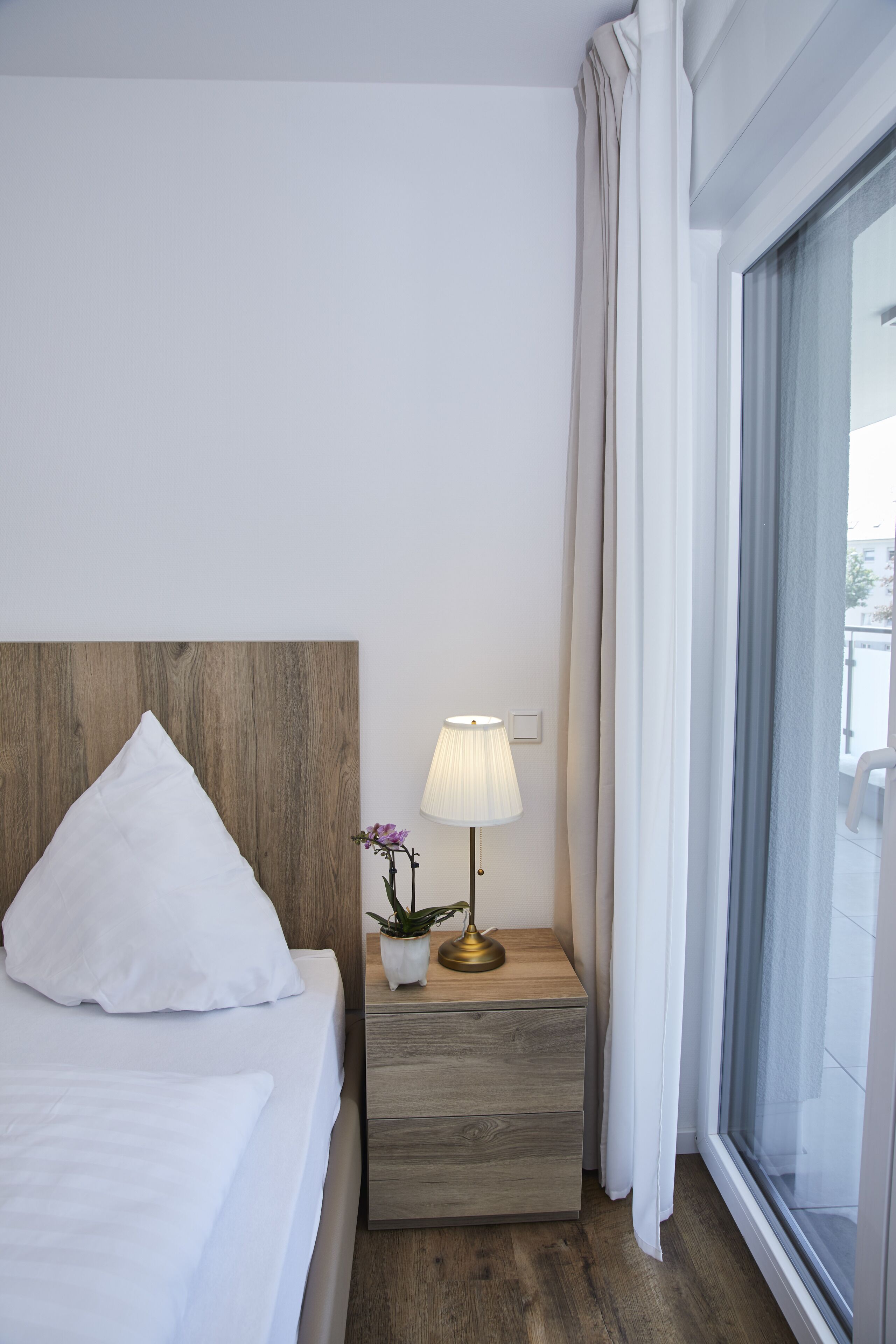 Een hotelkamer met een houten hoofdeinde, een wit bed, een nachtkastje met een lamp en uitzicht door een groot raam.