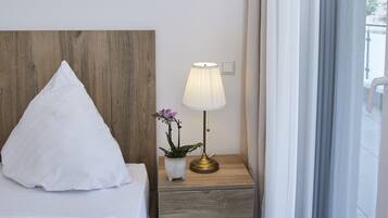 Een hotelkamer met een houten hoofdeinde, een wit bed, een nachtkastje met een lamp en uitzicht door een groot raam.