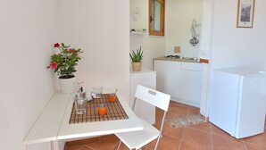 A compact kitchen with a white refrigerator, a small sink, and a window above the sink. There is a table with a placemat, a potted plant, and two orange cups.