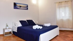 A neatly made bed with a blue bedspread and white pillows, a small bedside table with a plant, and a painting of a sunset on the wall.