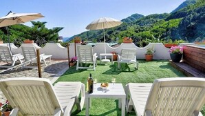 A rooftop terrace with white chairs, tables, and umbrellas, featuring a view of lush green hills.
