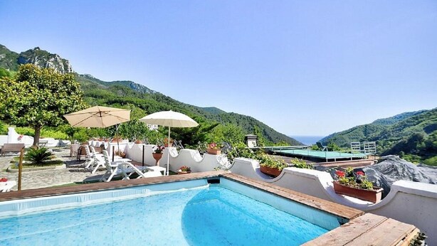 A pool area with lounge chairs and umbrellas, surrounded by greenery and mountains.
