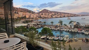 A balcony with a view of a marina, boats, and a cityscape with hills in the background. - Lion Gate Hotel (Sarandë)