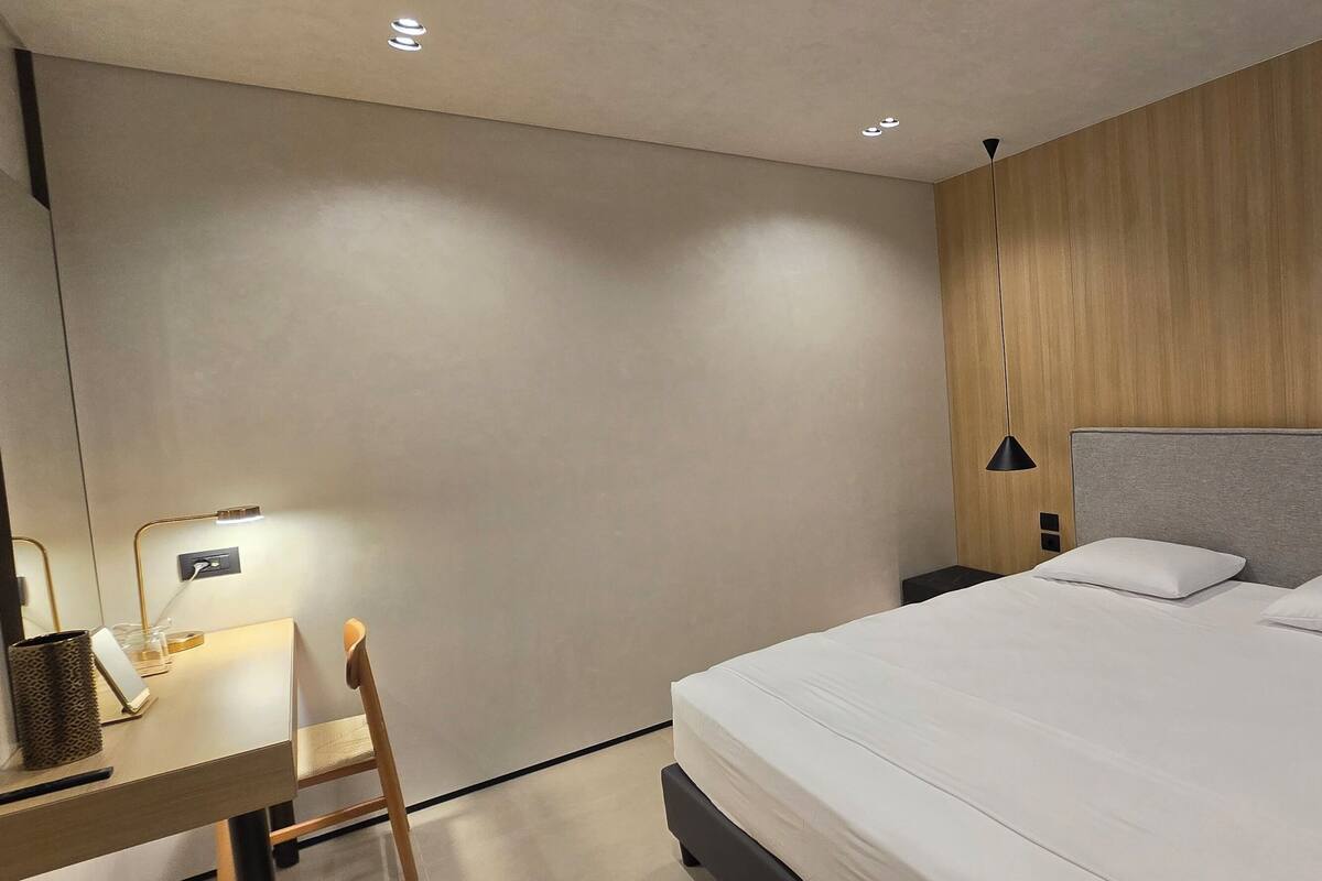 A modern hotel room with a large bed, two bedside tables with lamps, a desk with a chair, and a small table with a lamp.