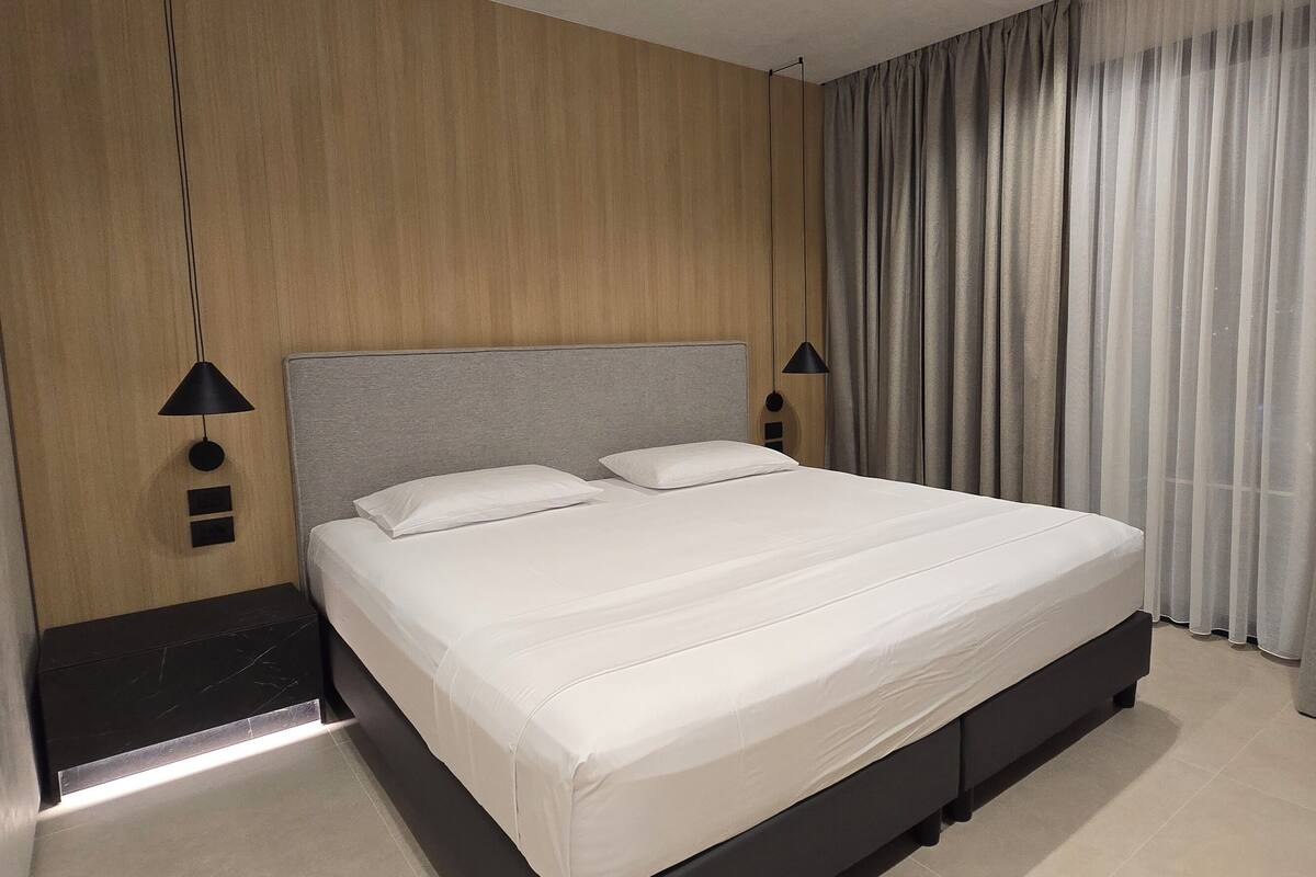 A neatly made bed with a grey headboard, a black nightstand, and a wall-mounted lamp.