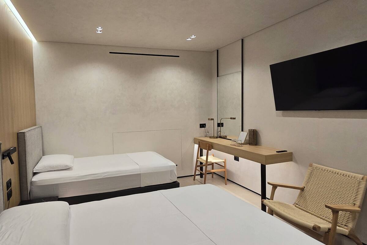 A modern hotel room with a large bed, a smaller bed, a desk with a chair, a mirror, and a TV mounted on the wall.