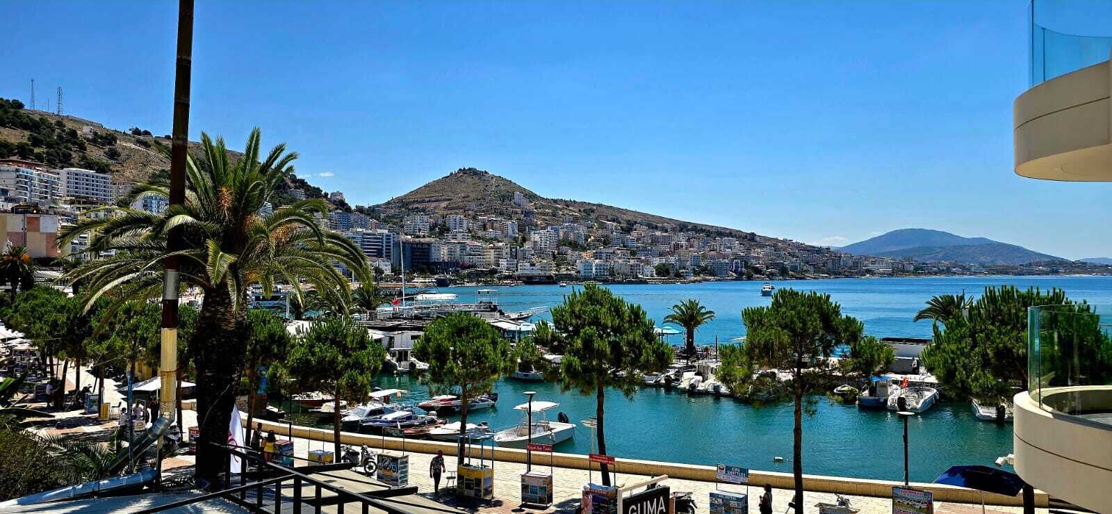 A coastal marina with boats, a clear blue sky, and a mountainous backdrop.