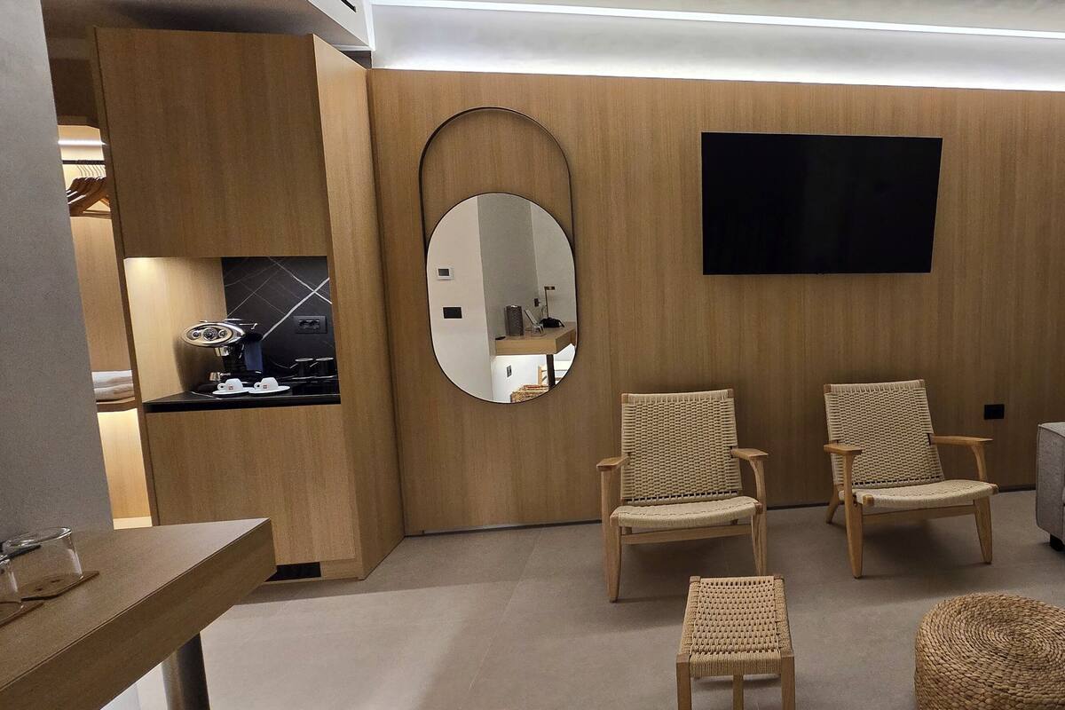 A modern hotel room with a wooden wall, a flat-screen TV, two wicker chairs, a grey sofa, and a small table with a mirror.