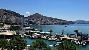 A marina with boats docked, a beach area with umbrellas, and a cityscape with buildings and a mountain in the background. - Lion Gate Hotel (Sarandë)