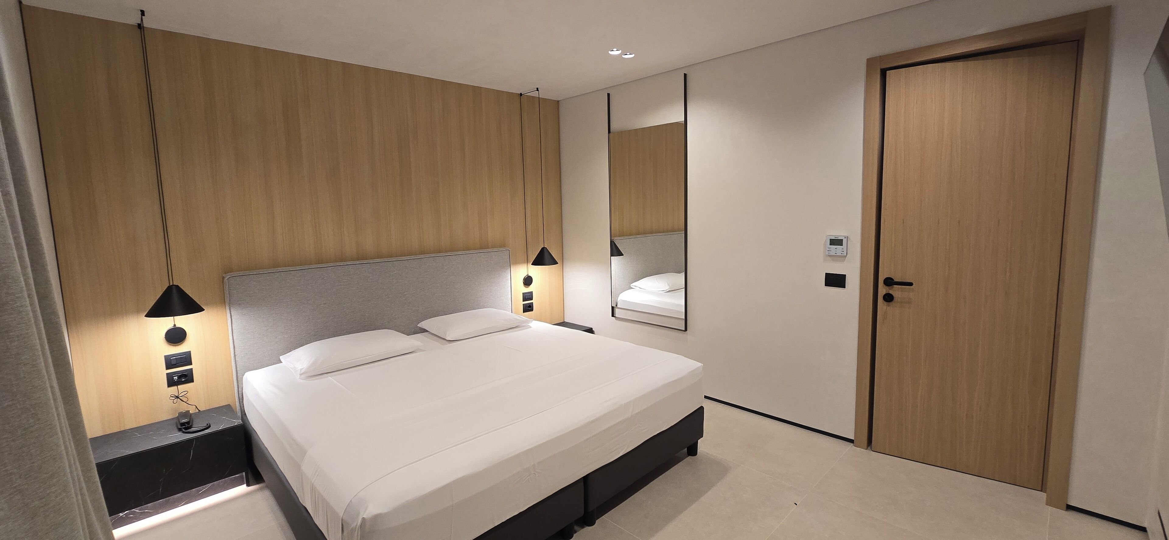 A modern hotel room with a large bed, bedside lamps, a nightstand, a mirror, and a wooden door.