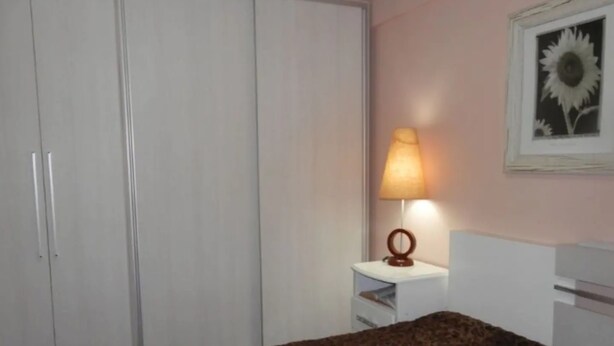 A bedroom with a bed, a wardrobe, a nightstand with a lamp, and a framed picture on the wall. - Apto Praia Central Camburiu (Balneário Camboriú)