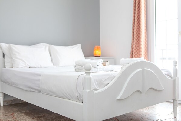 A white bed with a headboard, a nightstand with a candle, and a window with curtains.