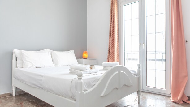 A white bed with pillows and a nightstand with a lamp in a room with a large window and a pink curtain.