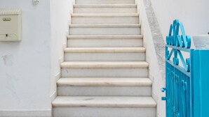 A staircase with white steps leading upwards, a blue metal railing on the right, and a white wall with a potted plant at the top.