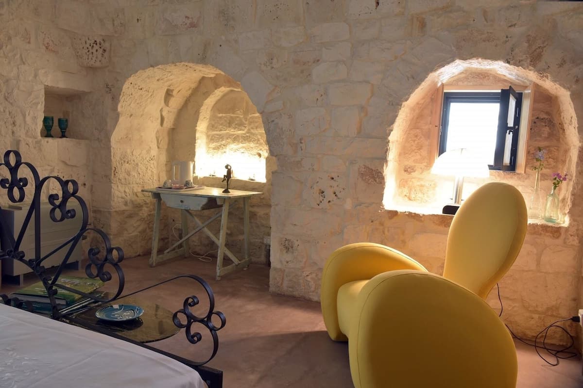 Una habitación rústica con paredes de piedra, una pequeña mesa con una silla, un ventanal con vistas y una cama con colcha blanca.