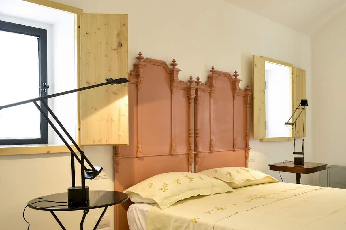 Un dormitorio con cabecera de madera, una cama con ropa de cama floral, una mesita de noche con lámpara y una ventana con persiana de madera.
