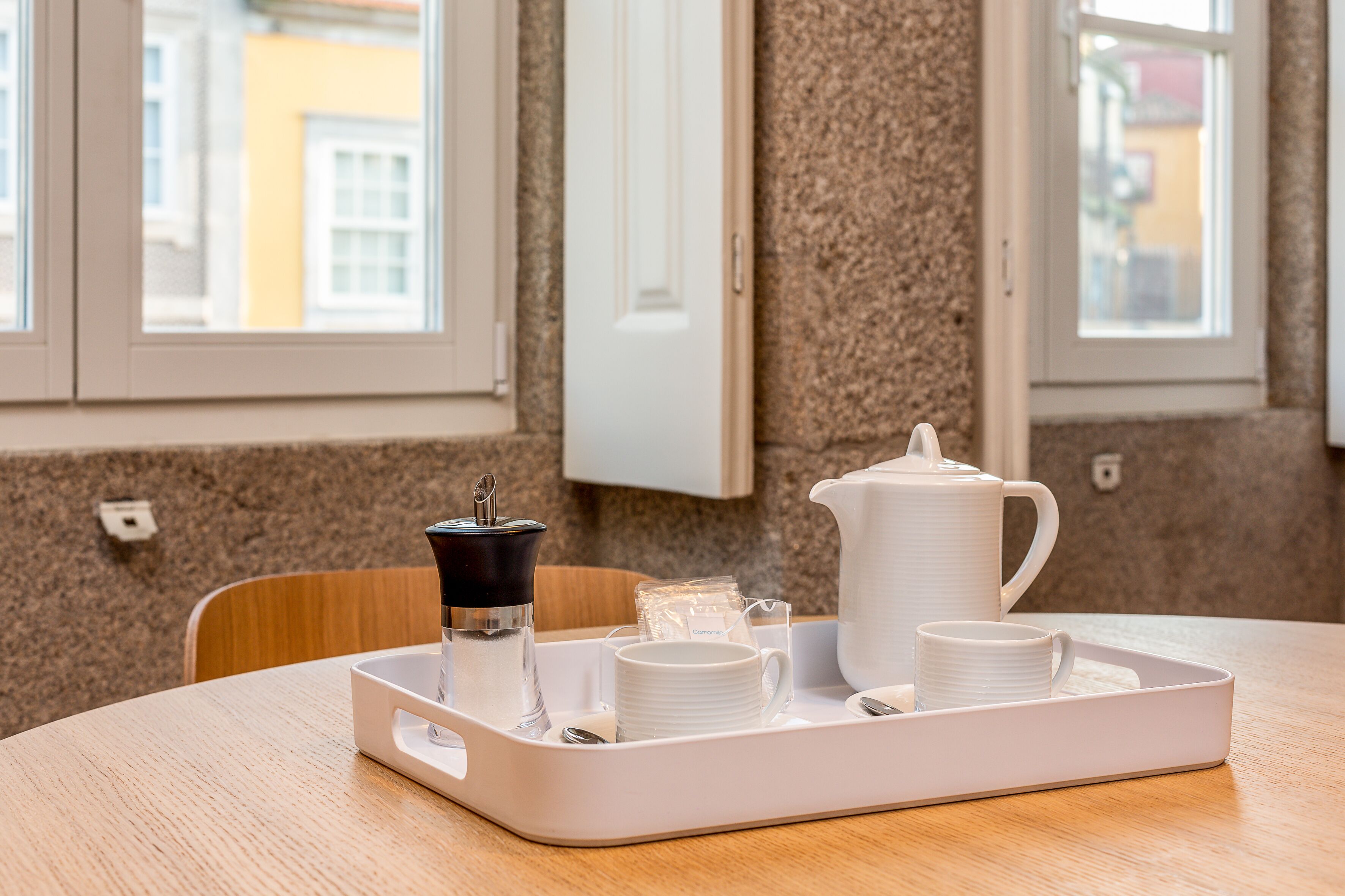 Ein Frühstücksbrett mit Kaffeemaschine, Tassen und einer Kanne auf einem Holztisch in einem Zimmer mit Fenster und Tür.