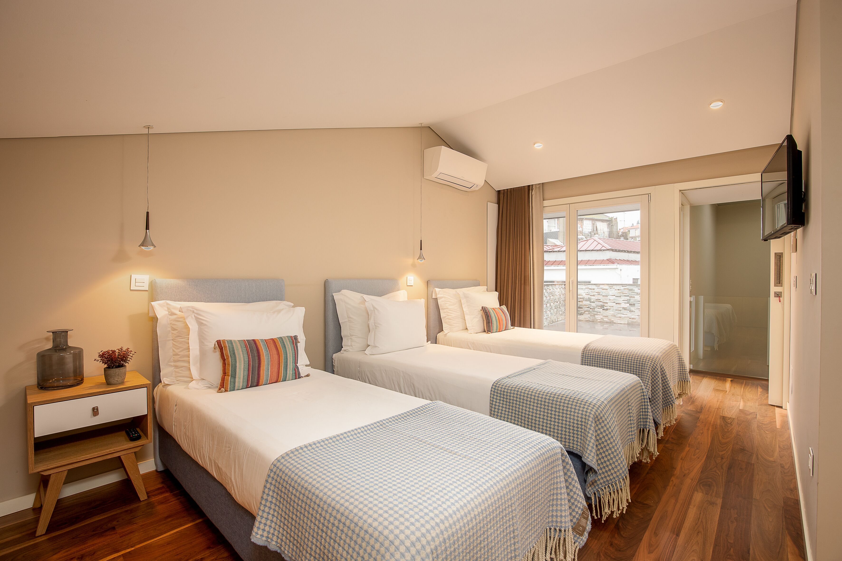 A hotel room with three beds, a bedside table with a lamp, an air conditioner, and a television mounted on the wall.