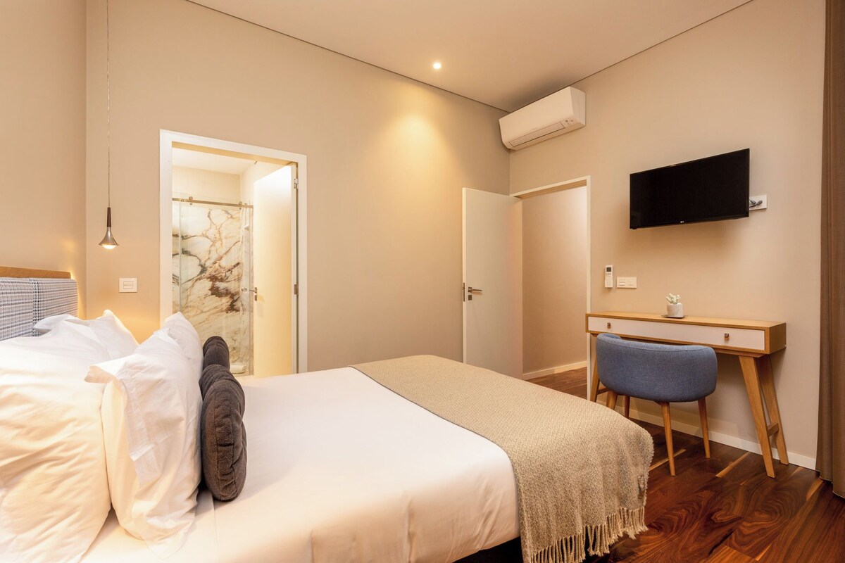 A bedroom with a bed, a desk, a chair, a TV, and an air conditioning unit.