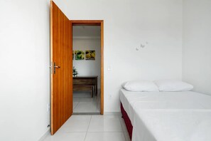 Un dormitorio con puerta de madera, una cama con ropa blanca, un escritorio pequeño con una silla y una estantería en la pared.