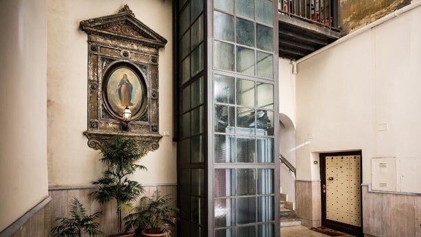A lobby with a large glass door, a framed artwork above a potted plant, and a staircase leading to an upper level.