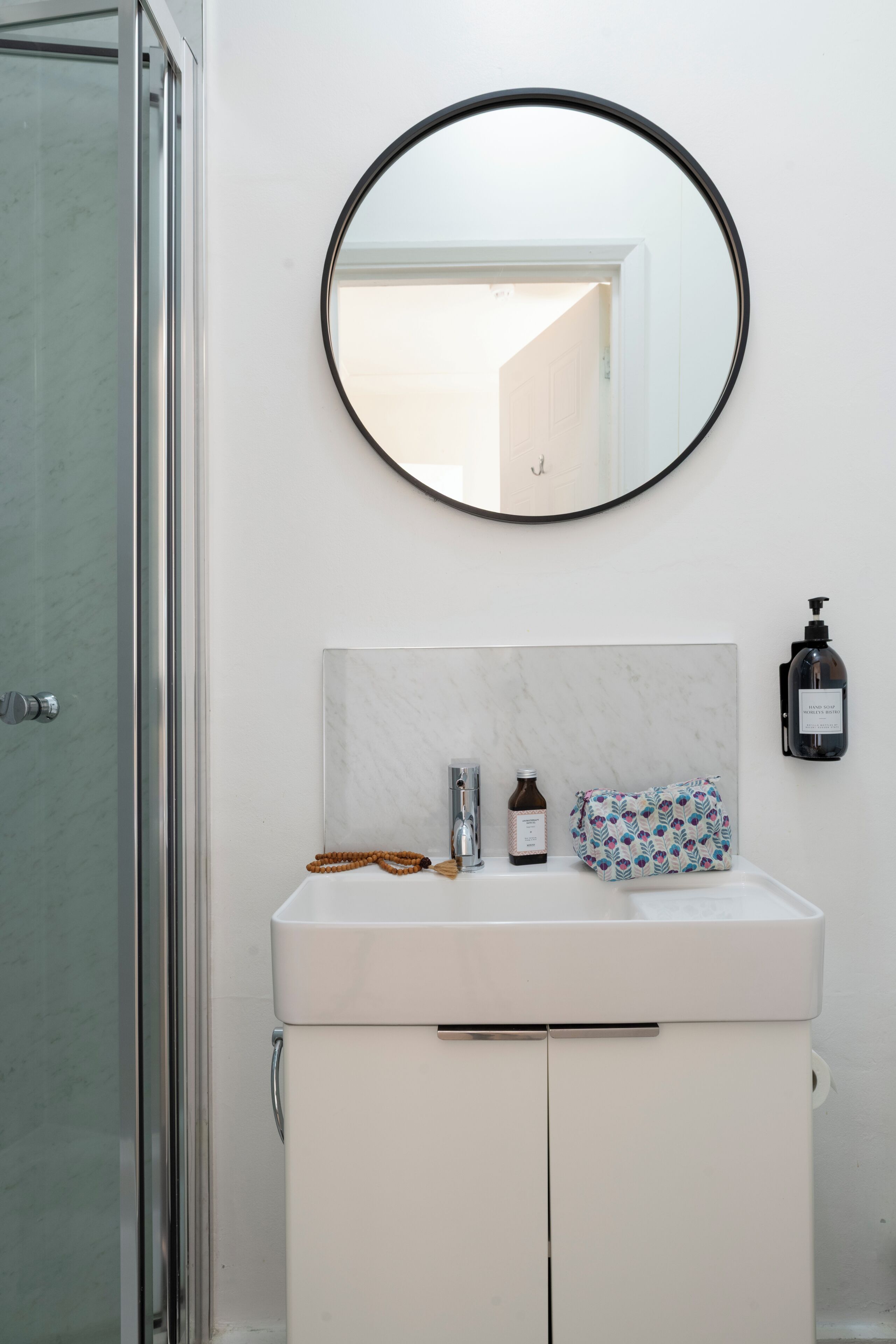 Un baño con un lavamanos blanco, un espejo redondo, un cabezal de ducha de mano y una botella de champú.