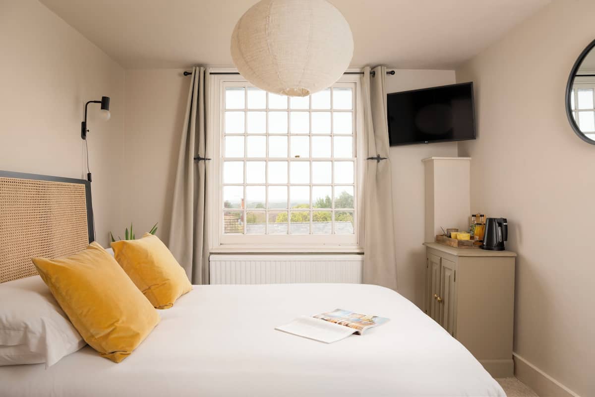 A bedroom with a bed, two pillows, a bedside table with a kettle, a TV, and a window with curtains.