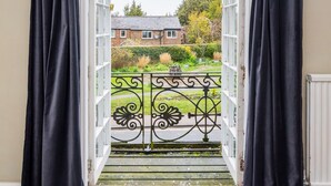 Une porte surmontée de rideaux noirs, qui donne sur un balcon doté d’une balustrade blanche et offrant une vue sur une maison et un jardin.
