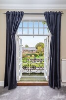 Une porte surmontée de rideaux noirs, qui donne sur un balcon doté d’une balustrade blanche et offrant une vue sur une maison et un jardin.