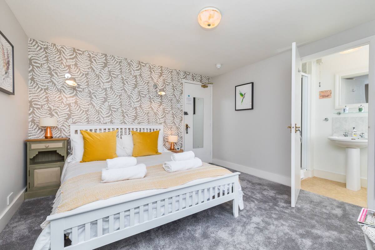 A bedroom with a white bed, a patterned wallpaper, a nightstand with a lamp, and a bathroom visible through an open door.