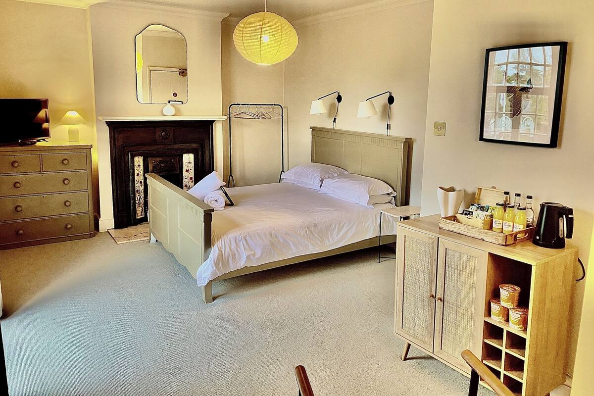 A bedroom with a bed, a dresser, a mirror, a hanging light, a fireplace, and a cabinet with items on top.
