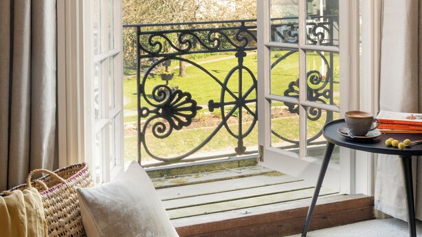 Une pièce avec une grande fenêtre, donnant sur un jardin, une petite table avec une tasse, et un banc avec des coussins.
