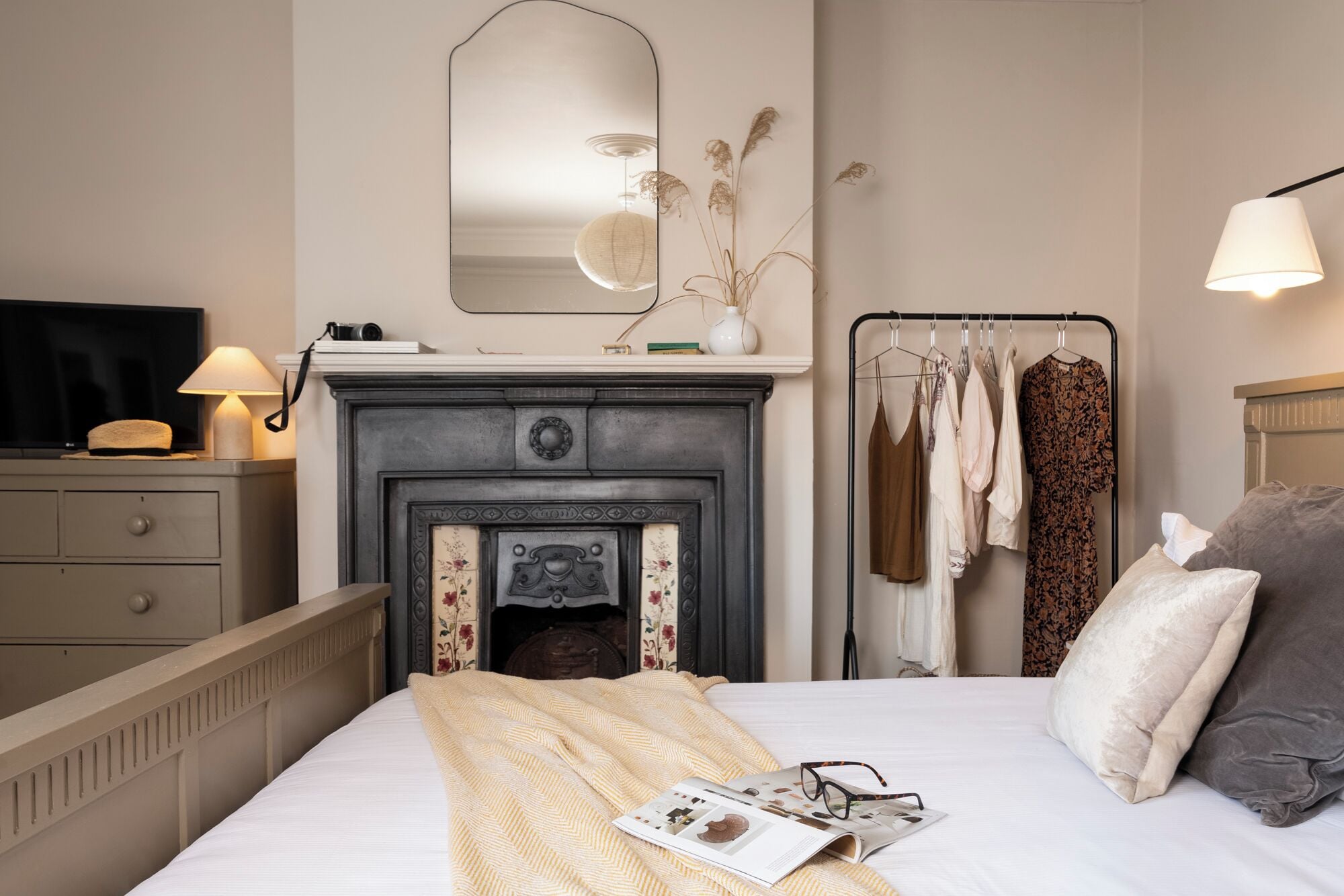 A bedroom with a bed, a dresser, a mirror, a fireplace, a clothing rack, and a lamp.