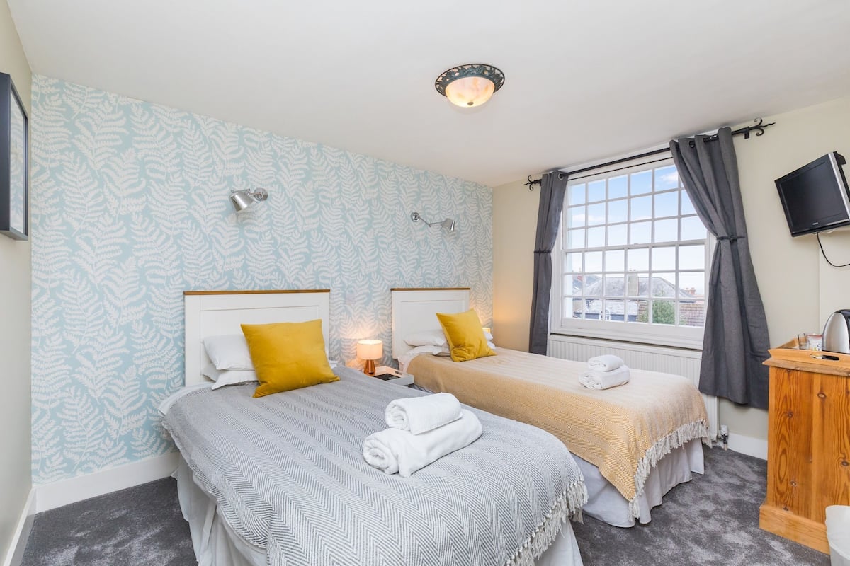 A hotel room with two beds, a TV, a window with curtains, a wooden cabinet, and a patterned wallpaper.