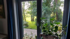Una ventana con vista a un jardín, un taburete de madera y un florero de metal con plantas.