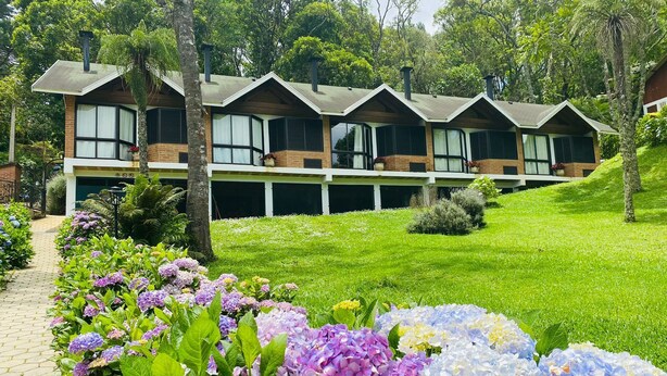 A row of modern, multi-story buildings with large windows and a flat roof, surrounded by lush greenery and a variety of flowers. - Terrazzo dos Alpes (Camanducaia)