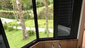A modern bathroom with a large bathtub, wooden paneling, and a view of a lush garden through large windows. - Terrazzo dos Alpes (Camanducaia)