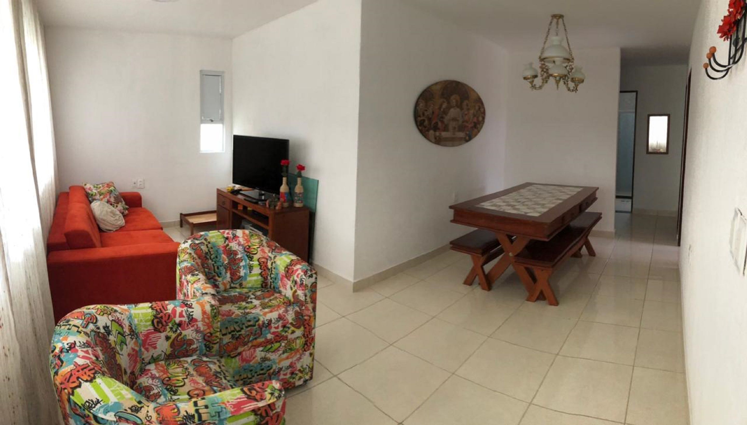 Uma sala de estar com um sofá vermelho, uma poltrona colorida, uma televisão e uma mesa de jantar com bancos.
