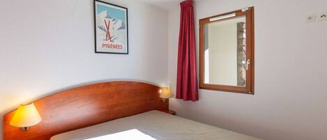 Une chambre à coucher avec un lit, une table de chevet, une lampe, une fenêtre avec un rideau et une affiche accrochée au mur.