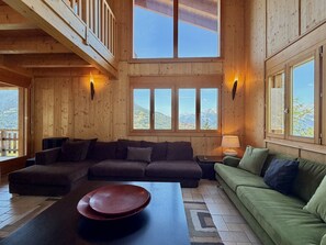 A wooden interior with a sectional sofa, a green couch, and a large window offering a mountain view.