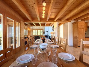 A wooden dining room with a long table set for a meal, wooden beams on the ceiling, and large windows.